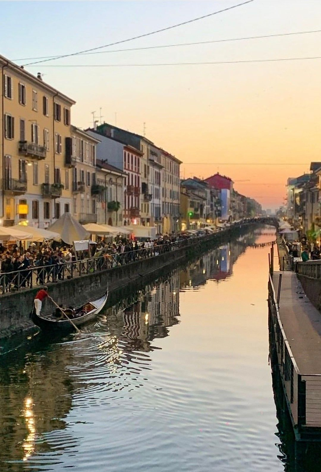 The Navigli canals in Milan at dusk, with lights reflecting on the dark water and people sitting along the banks.