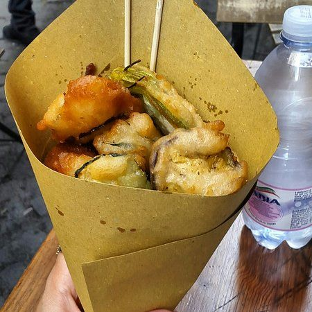 A paper cone of cuoppo overflowing with golden fried seafood and zucchini blossoms, held over the cobblestones of a Neapolitan market street.