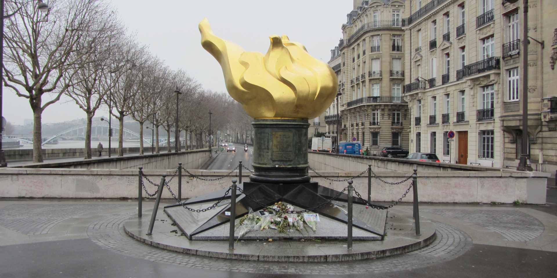 The golden Flame of Liberty memorial at the Pont de l'Alma in Paris, with fresh flowers and handwritten messages left by mourners at its base.
